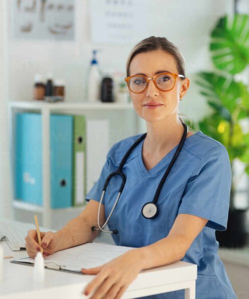 Portrait of a beautiful female doctor with eyeglasses, sitting at the desk in a doctor's office.